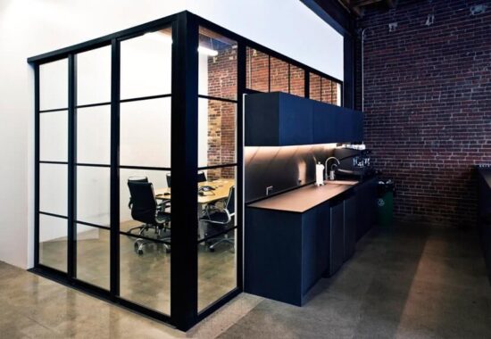 A modern office kitchenette adjacent to a glass-walled meeting room with exposed brick walls and interior glass doors.