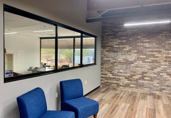 Modern office waiting area with blue chairs, brick accent wall, and a view into the adjacent office through glass doors.