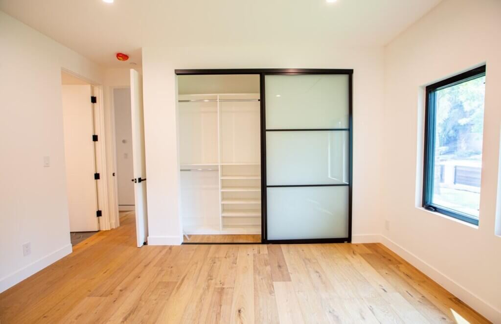 A bright, modern room with wooden flooring, featuring a sliding frosted glass interior door leading to a closet and a view of a window on the side.
