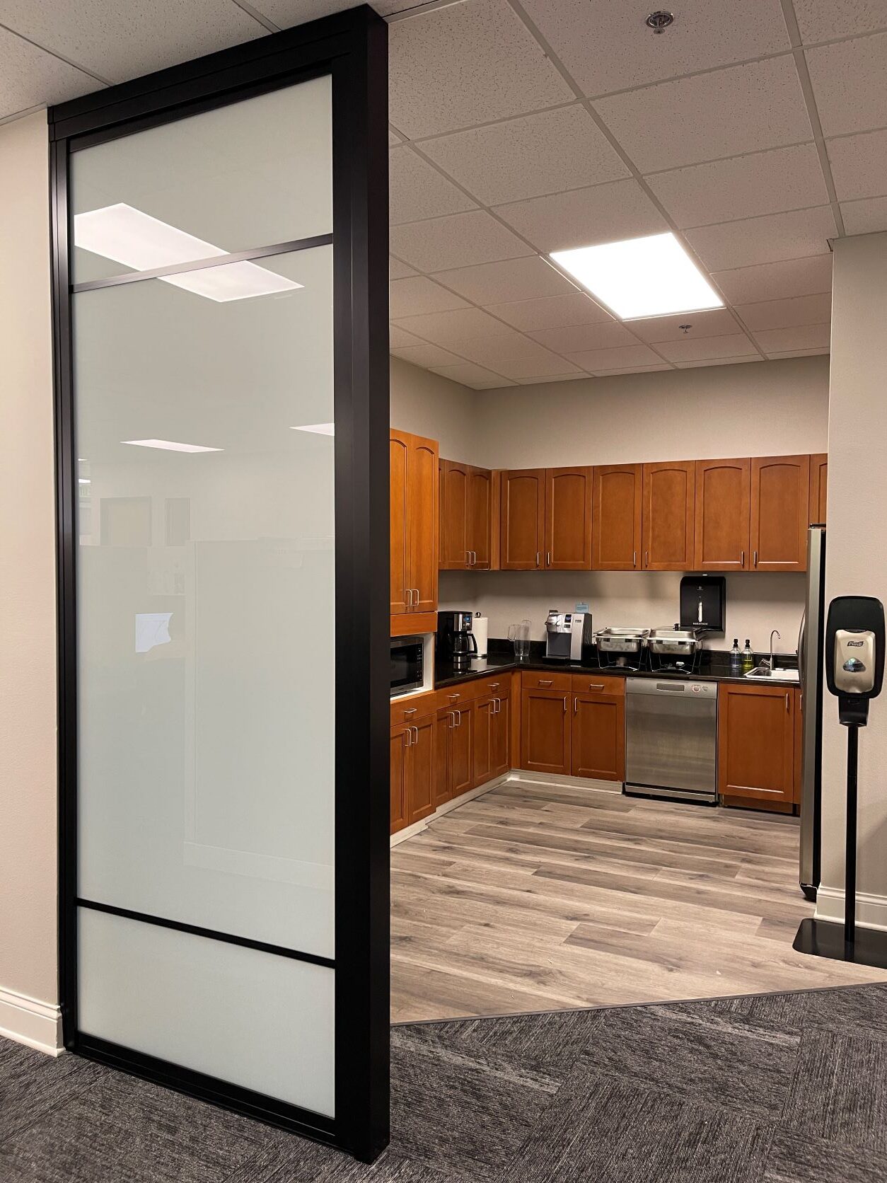 A modern office kitchen viewed through a glass door with a hand sanitizer dispenser by the entrance, featuring a fix panel.