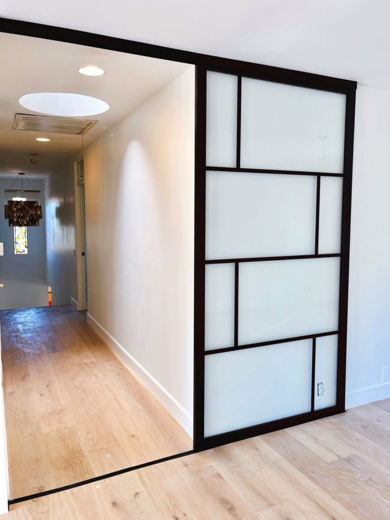 A modern interior with a large, sliding wall panel in a black frame, opening into a hallway with wooden flooring and a pendant light.
