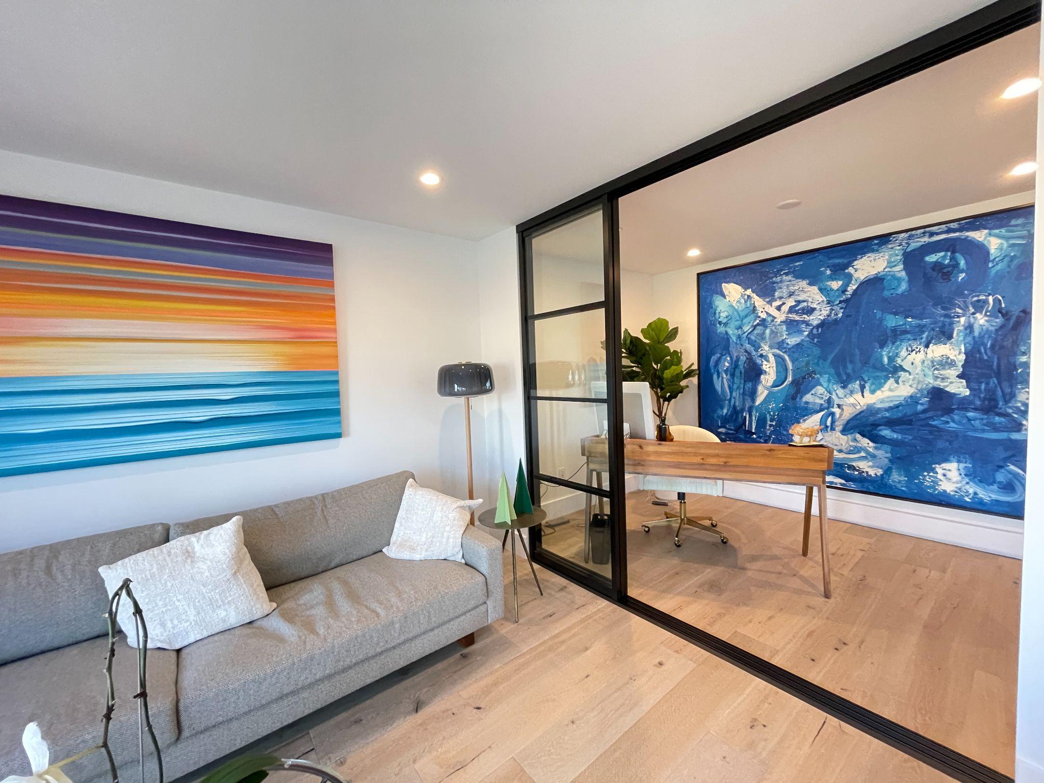 Interior sliding glass door separating an office from a living room with large artwork on the walls.