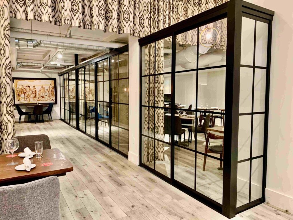 Modern restaurant interior with light wood flooring, glass partition walls, assorted chairs, and set dining tables; patterned curtains hang above the glass panels.