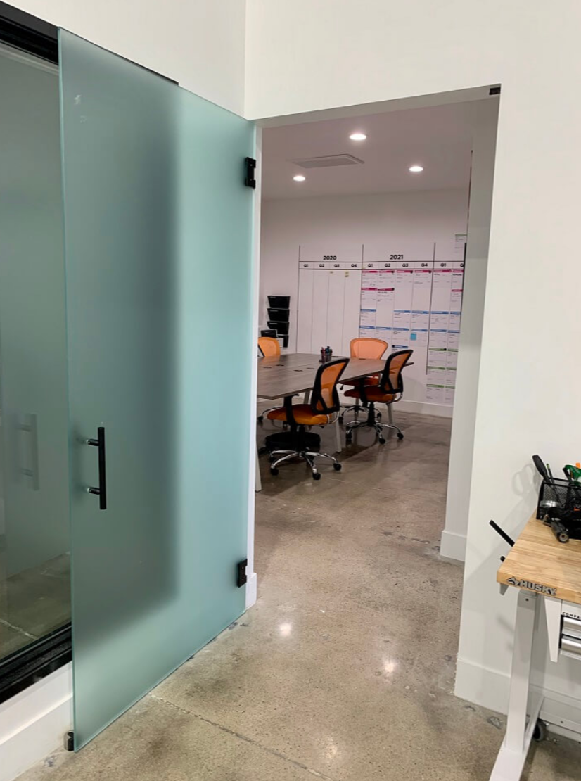 Office space with glass sliding door, several orange office chairs around a table, and a wall calendar with colored notes in the background.