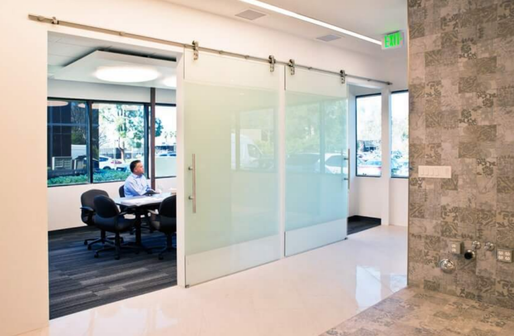 A man sits alone at a conference table in a modern office behind large frosted glass sliding doors, with windows showing trees and parked cars outside.