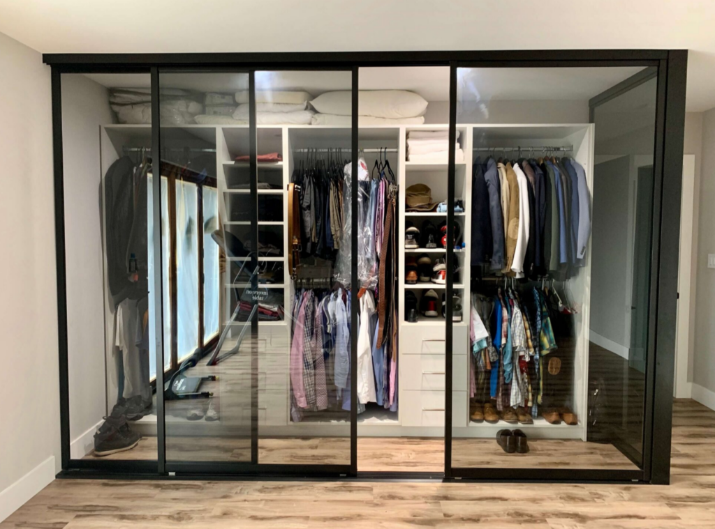 A modern wardrobe with glass sliding doors, organized with folded clothes, hanging shirts, jackets, pants, shelves for shoes, and storage boxes on the top shelf inside a bright room with wood flooring.